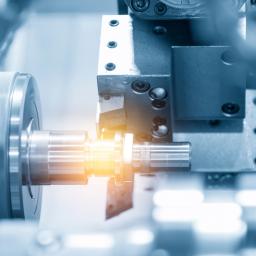 Close-up of a CNC lathe turning a metal shaft while a cutting tool machines the surface, with bright light reflecting off the workpiece.