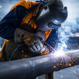 Welder in protective gear fusing a metal pipe, with bright sparks and blue-tinted smoke in an industrial workshop.