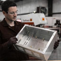 Man in black overalls inspecting a custom sheet metal enclosure in a UK fabrication workshop, holding a stainless steel housing with cut-outs and mounting points.