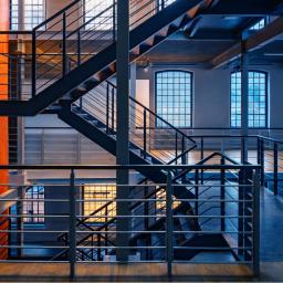 Modern industrial interior with multi-level steel staircases and railings, large grid windows, and a bold orange feature wall, styled with cool blue and warm orange lighting for a contemporary engineered look