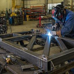 Alt text: A fabricator welds a large steel frame assembly on a workbench inside an industrial workshop, with sparks visible and tools and machinery in the background.