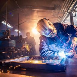 Welder fabricating a metal assembly on a workbench with bright blue arc light and orange sparks, set in a busy industrial workshop with machinery and warm backlighting.