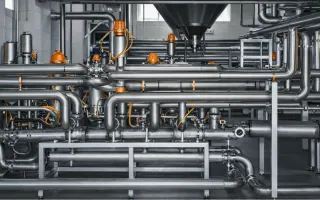 Complex stainless steel industrial piping system with bright orange valves and cables, arranged across a clean, modern processing facility.