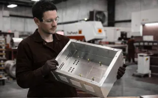 Man in black overalls inspecting a custom sheet metal enclosure in a UK fabrication workshop, holding a stainless steel housing with cut-outs and mounting points.
