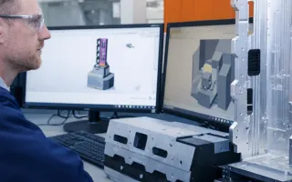 An engineer reviews CAD models on two monitors beside machined metal parts, with orange panels in the background.