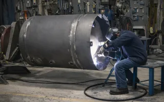 A fabricator sits on a blue metal stool welding inside the open end of a large cylindrical steel vessel in a workshop. Bright white welding light and small sparks illuminate the vessel interior, while tools, cables, and industrial equipment fill the background.