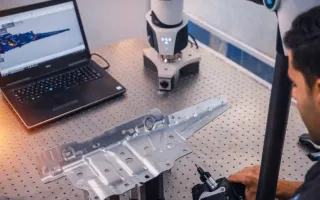 Engineer using a portable CMM arm to inspect a metal component on a perforated workbench, with a laptop displaying a CAD model, styled with subtle blue and orange lighting tones.