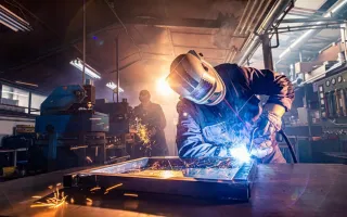 Welder fabricating a metal assembly on a workbench with bright blue arc light and orange sparks, set in a busy industrial workshop with machinery and warm backlighting.