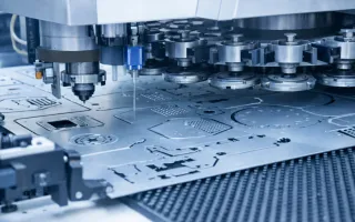 A CNC punching machine processes a metal sheet with cut-outs and perforations, shown in a flipped view with a light blue tint.