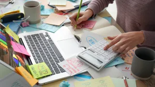 Cluttered desk showing manual sourcing and supplier research with notes, calculations, and paperwork