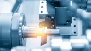Close-up of a CNC lathe turning a metal shaft while a cutting tool machines the surface, with bright light reflecting off the workpiece.