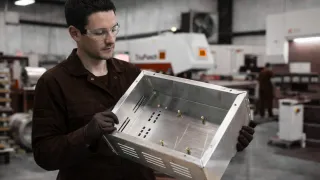 Man in black overalls inspecting a custom sheet metal enclosure in a UK fabrication workshop, holding a stainless steel housing with cut-outs and mounting points.