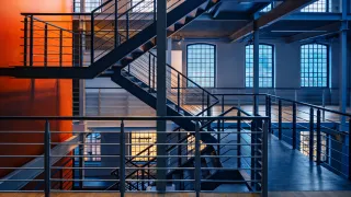 Modern industrial interior with multi-level steel staircases and railings, large grid windows, and a bold orange feature wall, styled with cool blue and warm orange lighting for a contemporary engineered look