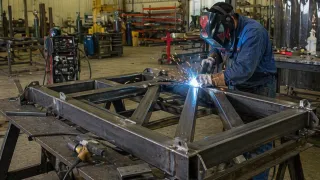Alt text: A fabricator welds a large steel frame assembly on a workbench inside an industrial workshop, with sparks visible and tools and machinery in the background.