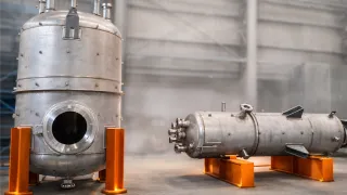 Two large stainless steel pressure vessels stand in a factory, with orange support sections and a softly blurred workshop background.