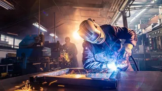 Welder fabricating a metal assembly on a workbench with bright blue arc light and orange sparks, set in a busy industrial workshop with machinery and warm backlighting.