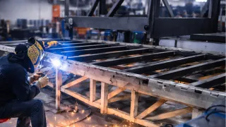 Welder seated on a stool fabricating a large steel frame in a workshop, with bright blue arc light and orange sparks illuminating the industrial environment.